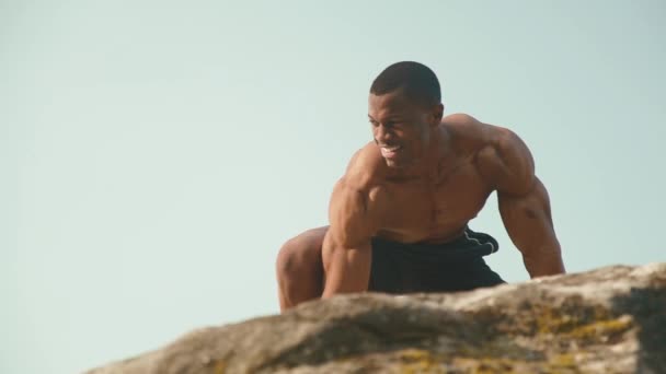 Närbild av stilig svart afrikansk amerikansk man bodybuilder med naken torso poserar på Rocky Mountain Peak med blå himmel bakgrund. Wild Nature Style. — Stockvideo