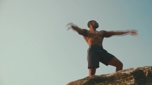 Atleta afro-americano com tronco nu posando na rocha e levantando as mãos para o céu — Vídeo de Stock