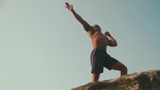 Handsome smiling african american athlete with naked torso posing on the rock and raising hands to the sky — Stock Video