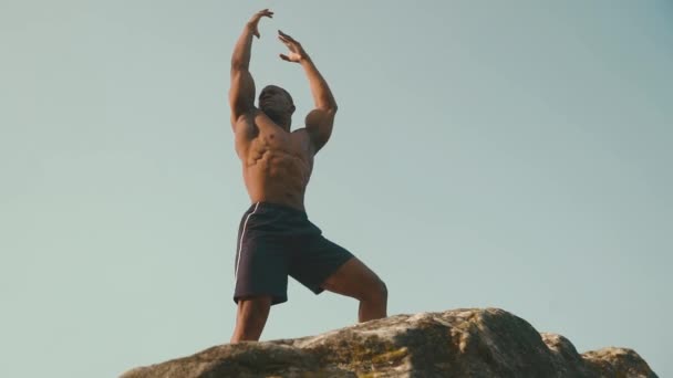Schöner lächelnder afrikanisch-amerikanischer Bodybuilder mit nacktem Oberkörper, der auf dem Felsen posiert und die Hände in den Himmel reckt. Harmonie zwischen Mensch und Natur — Stockvideo