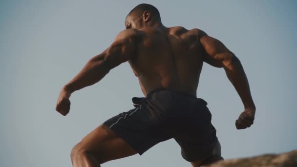 Vue de dos sur l'homme sportif afro-américain avec torse nu posant sur la roche et montrant les muscles. Fond ciel bleu clair — Video