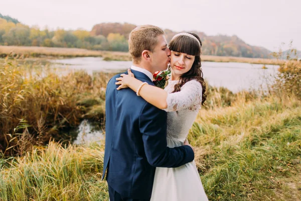 Portret van jonge jonggehuwde paar buitenshuis. Bruidegom kussen zijn charmante bruid in Wang over riverside — Stockfoto