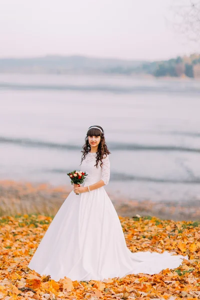 Belle jeune mariée brune innocente en magnifique robe blanche se tient sur des feuilles tombées au bord de la rivière — Photo