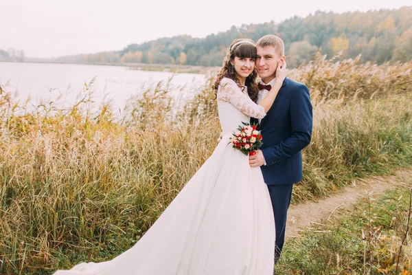 Retrato de una hermosa pareja joven recién casada al aire libre. Feliz novio sosteniendo a su novia en la orilla del río — Foto de Stock