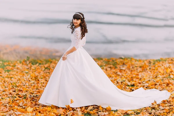 Hermosa inocente joven novia morena en vestido blanco magnífico se para en hojas caídas a orillas del río —  Fotos de Stock