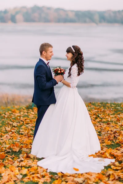 Jeune couple de jeunes mariés tenant la main sur le bord du lac d'automne plein de feuilles d'orange — Photo