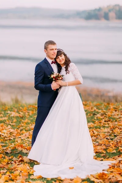 Joven pareja nupcial recién casada sosteniéndose mutuamente en la orilla del lago de otoño llena de hojas naranjas —  Fotos de Stock