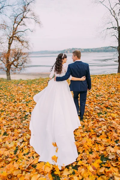 Heureux jeunes mariés marchant sur le bord du lac d'automne plein de feuilles d'orange — Photo