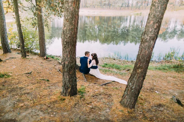 Noivo feliz e sua encantadora nova esposa segurando um ao outro enquanto sentado na margem do lago da floresta — Fotografia de Stock