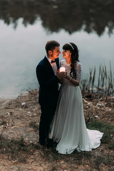 Brudgummen och hans charmiga nya fru med brinnande ljus på stranden vid skogstjärn på kvällen — Stockfoto