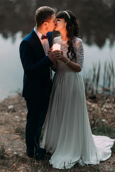 Groom e sua encantadora nova esposa se beijando na margem do lago da floresta à noite — Fotografia de Stock