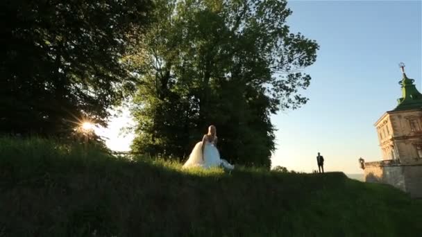 Joyeux jeune mariée blonde en robe blanche et marié en costume s'amuser et sauter près d'un vieux château médiéval — Video