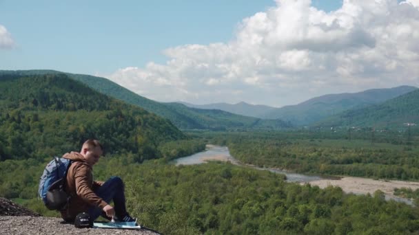 Joven hombre activo comprobando el terreno del paisaje con mapa en roca de montaña — Vídeo de stock