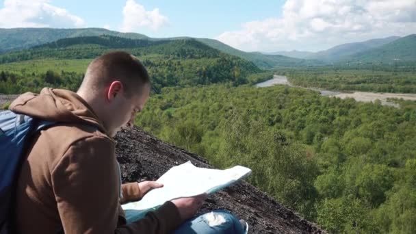 Joven hombre activo comprobando el terreno del paisaje con mapa y brújula mientras está sentado en un montículo rocoso — Vídeos de Stock