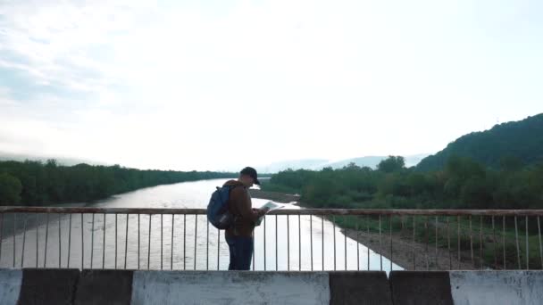 Unga manliga turist kontrollera sin väg på rutt med karta när man går på bron i bergen — Stockvideo