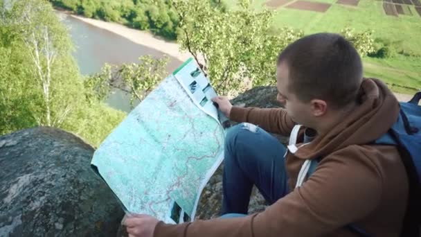 Unga turist man kolla sin rutt med karta medan du sitter på skuggad rock boulder med floden Visa nackdelen — Stockvideo