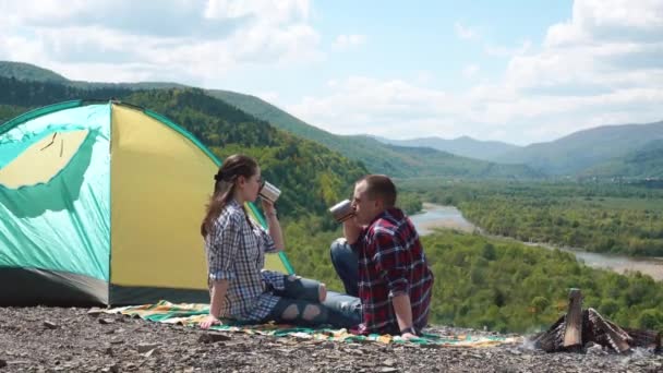 Jovem casal turístico bebe chá e beijos no local do acampamento com fogueira no topo rochoso da colina em montanhas verdes — Vídeo de Stock