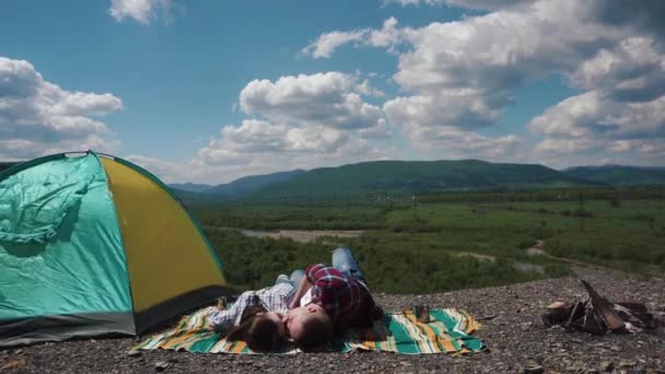 Mladí turistické pár líbání při odpočinku na dece v kempu backgrounded zelené hory a údolí pod modré oblohy jasno — Stock video