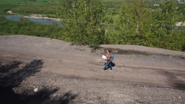 Jovem turista caminha na estrada de cascalho acima do vale do rio em montanhas verificando seu caminho com papel mapa — Vídeo de Stock