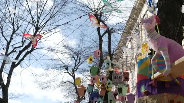 Fancy colorful toys and lanterns, decorated as birdhouses, swinging on leafless tree on european city street — Stock Video