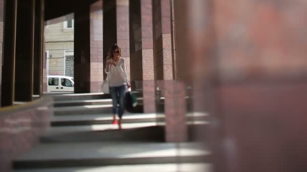 Приваблива ділова жінка з сумками, що ходять по місту і ховаються від ранкового сонця за колонами сучасної будівлі стилю — стокове відео