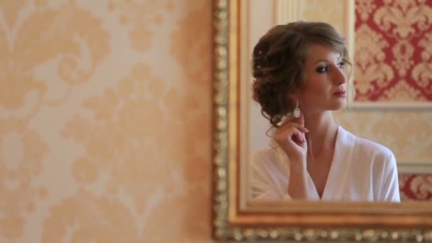 Bride in silk housecoat looking at mirror, trying on her wedding earrings at morning of the marriage day — Stock Video