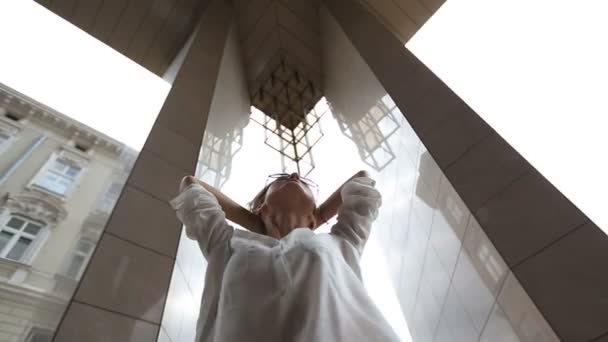 Attraktive junge lächelnde Frau in weißem Hemd mit zusammengerollten Ärmeln, die neben Marmorsäulen des modernen Stadtbaus steht und ihre Haare im Wind streift — Stockvideo