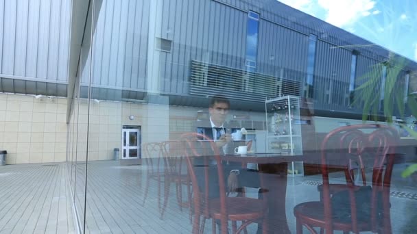 Деловой человек пьет кофе, сидит в ярком кафе. Вид из большого зеркального окна — стоковое видео