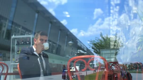 Hombre de negocios bebiendo café, sentado en la cafetería brillante con espejo de la pared de cristal exterior. Cielo azul nublado en reflexión — Vídeos de Stock