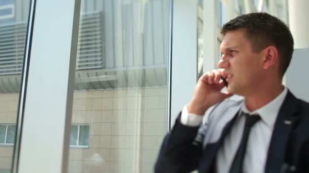 Bello giovane uomo d'affari ride mentre parla al telefono e guardando nella finestra dell'edificio moderno — Video Stock