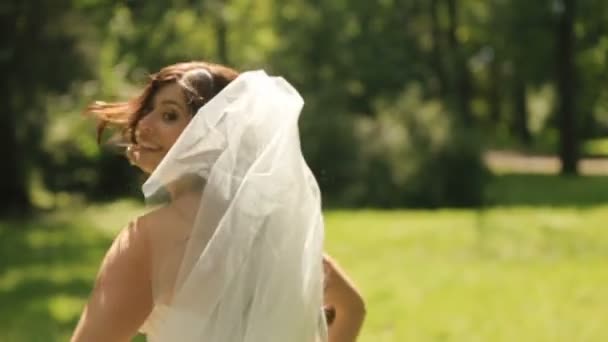 長いベールを持つ優秀白ドレスで素敵な豪華な花嫁は公園の芝生の上ふざけて逃げる — ストック動画