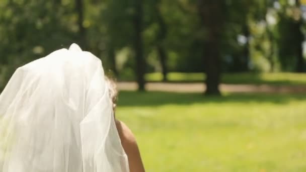 Wunderschöne Braut in vorzüglichem weißen Kleid mit langem Schleier läuft spielerisch auf dem Rasen im Park davon — Stockvideo