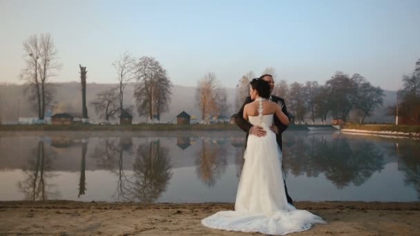 Dos recién casados felices abrazándose en la noche del día de la boda en el lago del complejo de montaña en la puesta del sol — Vídeos de Stock