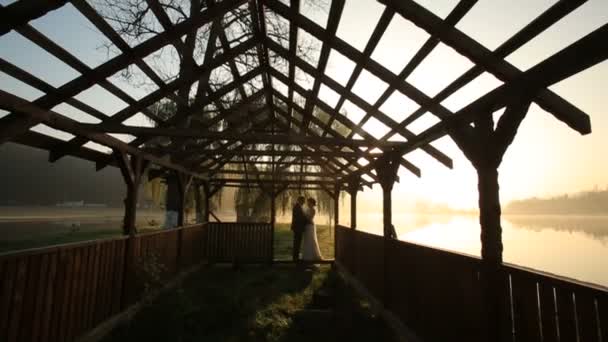 Glückliches frischvermähltes Paar, das sich am Abend des Hochzeitstages am Bergsee bei Sonnenuntergang in Strandnähe umarmt — Stockvideo