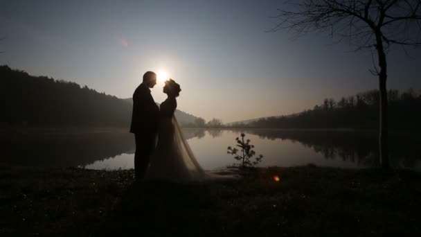 Glückliches frischvermähltes Paar, das sich am Abend des Hochzeitstages am Bergsee bei Sonnenuntergang in Strandnähe umarmt — Stockvideo