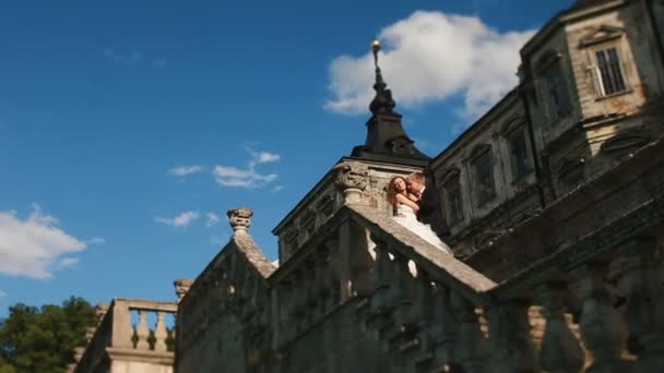 Loving groom tenderly kisses his beautiful bride while embracing her on the stairs of old castle. Magnificent blue sky with small clouds as background — Stock Video
