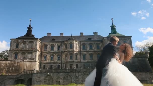 Loving groom tilts his beautiful bride and kissing her in neck. Magnificent antique castle under blue sky with small clouds as background — Stock Video