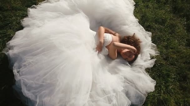 Чувственная молодая невеста в роскошном белом свадебном платье, лежащем на зеленом газоне и соблазнительно смотрящем в камеру — стоковое видео