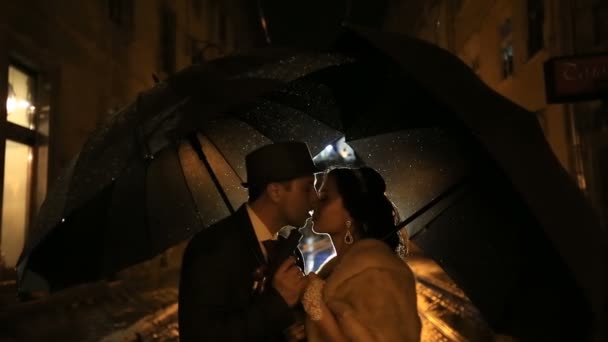 Beautiful noir-styled couple under rain kissing on night street at rain. Pair holding umbrellas, young man playing a gangster — Stock Video