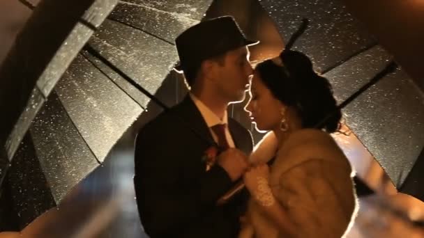 Прекрасная пара в стиле нуар целуется на ночной городской улице, прячется от дождя под зонтиками — стоковое видео