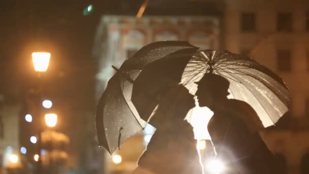 Romantic noir-styled couple under rain kissing on night city street, hiding from rain under umbrellas. City lights show pair silhouettes — Stock Video