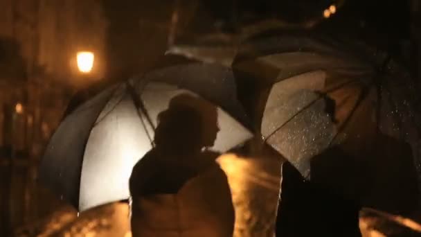 Belo casal caminha sob chuva na rua da cidade noturna, escondendo-se da chuva sob guarda-chuvas. luzes da cidade mostrar silhuetas par — Vídeo de Stock