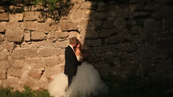 Amare sposo teneramente tiene la sua bella sposa mentre abbraccia la sua piedi vicino antico muro in rovina — Video Stock