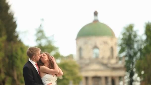 Αισθησιακό ακριβώς παντρεμένο ζευγάρι ευτυχώς αγκαλιάζοντας στο πάρκο με μακρινό παλαιά εκκλησία στο παρασκήνιο — Αρχείο Βίντεο