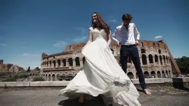 Свадебная пара в Риме танцует босиком на крыше возле Колизея, счастливый жених смотрит, как его невеста играет со своим длинным свадебным платьем. Медовый месяц в Италии, Европе — стоковое видео