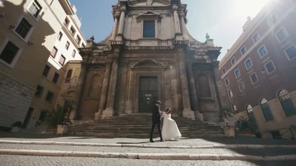 Bruiloft echtpaar in Rome wandelen voor San Nicola da Tolentino kerk. Stijlvolle bruidegom zachtjes bezit is van de hand van zijn prachtige bruid. Wittebroodsweken in Italië, Europa — Stockvideo