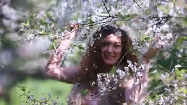 Young girl shaking a blossoming cherry branch in spring orchard and laughing when petals fall down like snowflakes on her curly hair — Stock Video