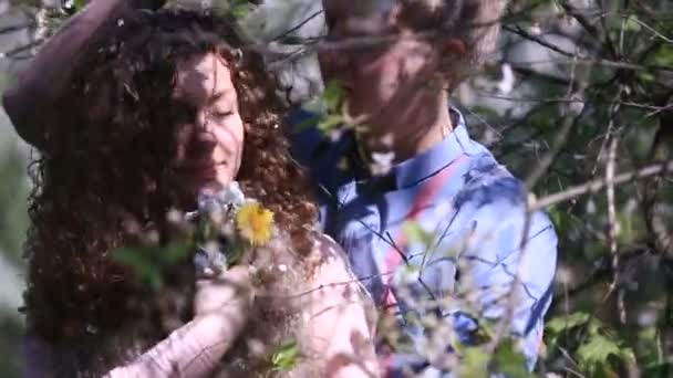 Ρομαντικό ζευγάρι στην άνθηση κεράσι οπωρώνα με πολλά λουλούδια. Νεαρός άνδρας αγκαλιάζει τη φίλη του και τα κατοικίδια ζώα σγουρά μαλλιά με λευκά πέταλα σε αυτό — Αρχείο Βίντεο