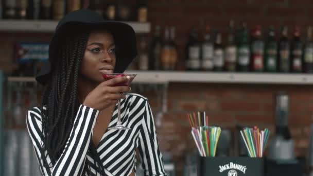 Primer plano de moda mujer afroamericana elegante en sombrero negro y vestido a rayas beber cóctel en el bar en el día caliente de verano — Vídeo de stock