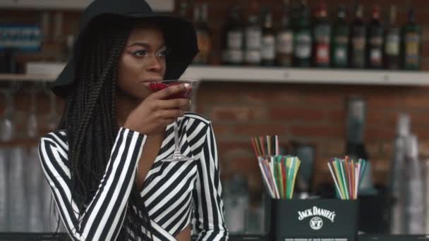 Sexy fashionable african american lady in black hat and striped vogue dress drinking cocktail in bar. Hot summer day refreshness — Stock Video